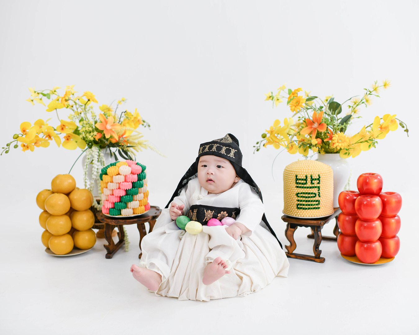 노란 꽃과 전통 백일상 앞에 앉은 한복 차림의 아기