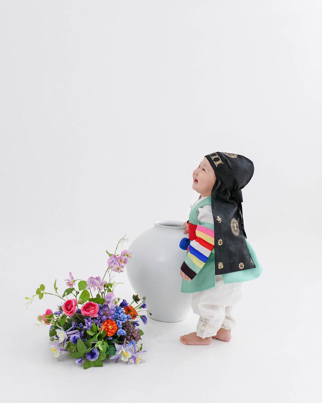 민트색 한복을 입고 꽃과 항아리 옆에 서서 웃는 아기