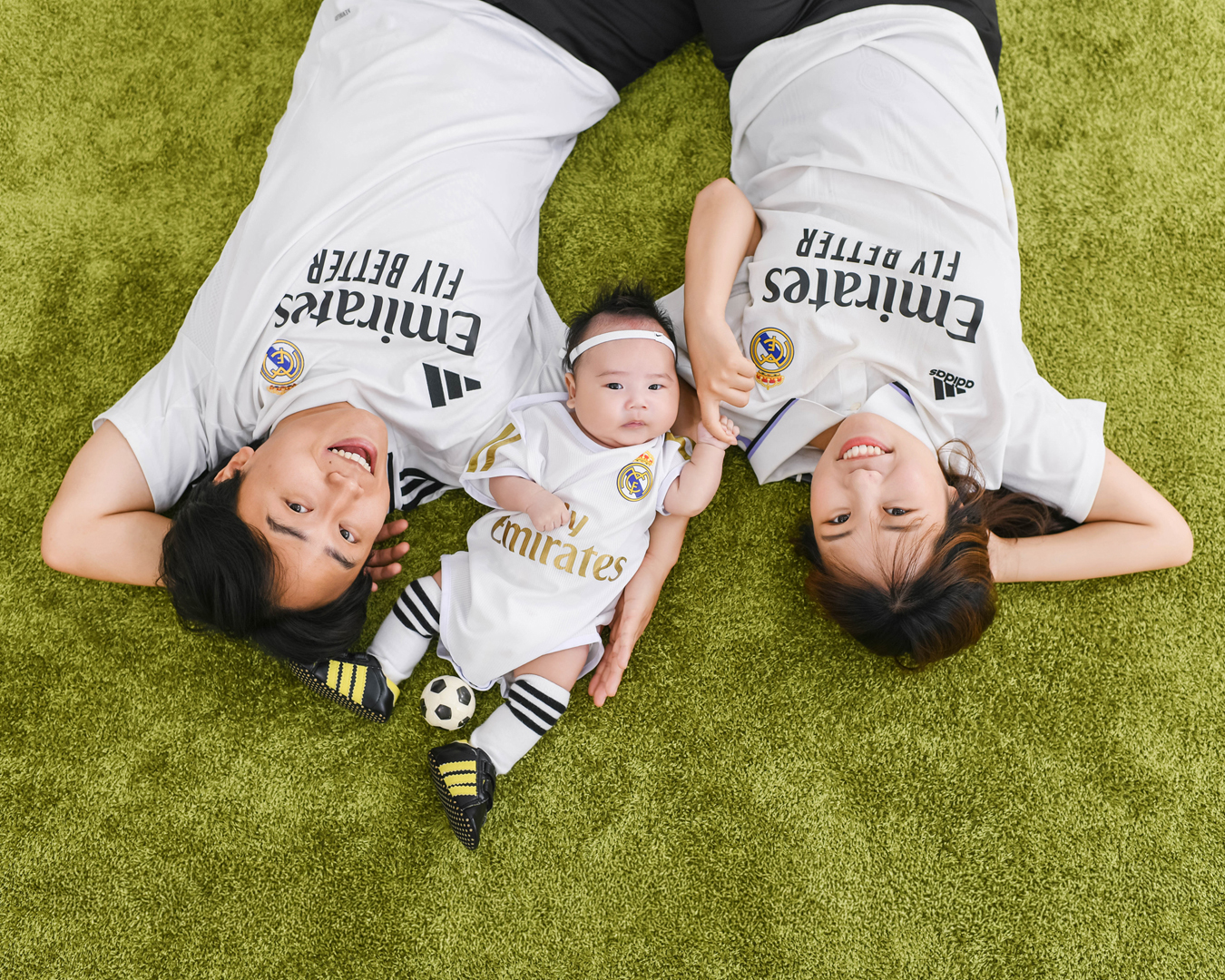 잔디 위에 같은 축구 유니폼을 입고 나란히 누운 아기와 부모